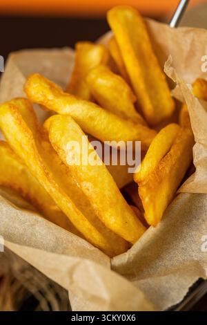 Frites dorées croustillantes dans un panier en papier sur une table sombre, gros plan avec un éclairage chaud Banque D'Images