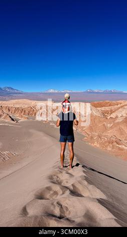 Photo verticale d'un touriste méconnaissable tenant son fils ou son tout-petit sur les épaules, tout en se tenant debout au sommet de la dune et en regardant Licancabur Banque D'Images