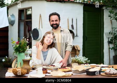 Homme d'âge moyen caucasien debout derrière une femme d'âge moyen caucasienne assise à la table extérieure, à la fois souriant et préparant la nourriture ensemble, abri rustique et outils de jardin en arrière-plan Banque D'Images