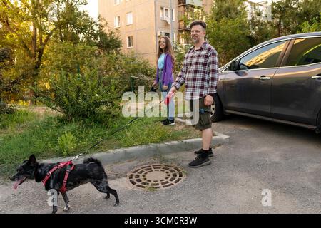 L'homme et sa fille sourient tout en promenant leur chien noir dans un harnais rouge près d'une voiture garée, tenant des laisses et profitant de la compagnie à l'extérieur dans un Banque D'Images