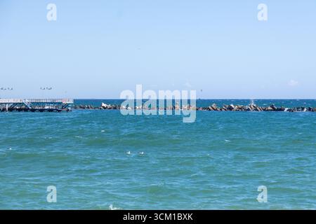Vue côtière des vagues roulant sur le rivage sablonneux près d'un brise-lames en pierre sous un ciel bleu clair Banque D'Images