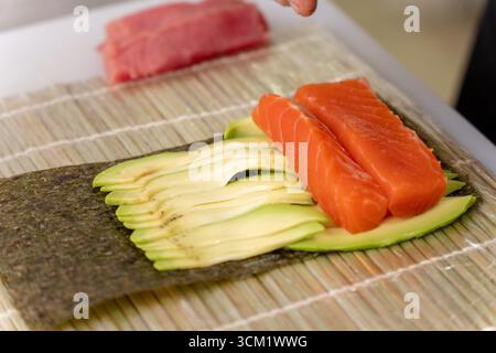 Rouleau de sushi fraîchement préparé avec du saumon, du thon et de l'avocat, prêt à être enroulé sur un tapis de bambou. Délicieuse cuisine maison. Banque D'Images