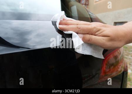 Une main polit délicatement une voiture noire élégante avec un tissu blanc, reflétant la lumière du soleil et les environs. Banque D'Images