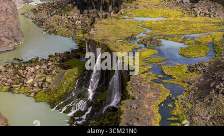Perspective aérienne d'une double cascade se jetant dans une rivière sinueuse, entourée de mousse verte, de roches volcaniques et d'eau turquoise contrastée à Icel Banque D'Images