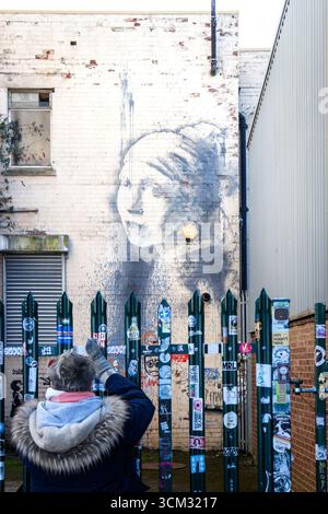 Une femme photographie une murale banksy inspirée par Painting Girl With a Pearl Earring sur un mur urbain derrière une clôture dans une ruelle de la ville de bristol Banque D'Images