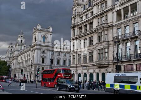 Une scène de rue à Londres avec une architecture grandiose, un bus à impériale rouge, un taxi noir et un van de police sous un ciel nuageux. Banque D'Images