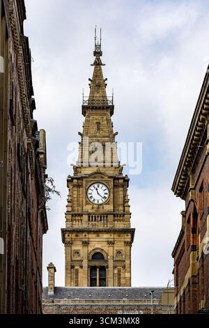 Une tour d'horloge en pierre avec des chiffres romains se dresse entre deux bâtiments en briques sous un ciel nuageux à Liverpool, Royaume-Uni. Banque D'Images
