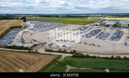 Vue aérienne d'un grand terrain de stockage extérieur de voitures, rempli de rangées de véhicules et entouré de champs et de bâtiments industriels sous un ciel nuageux à C. Banque D'Images