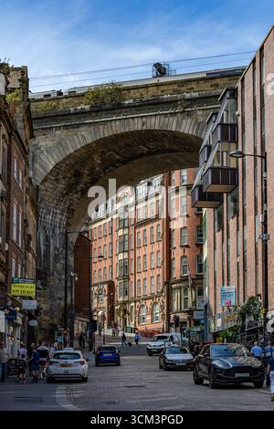 Un pont en arc de pierre enjambe une rue de la ville bordée de bâtiments en briques. Voitures et piétons sont présents dans la rue sous un ciel bleu à Newcastle upon Banque D'Images