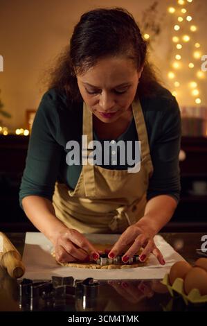 Une femme portant un tablier de chef prépare méticuleusement les biscuits des fêtes à l'aide d'emporte-pièce tout en étant entourée de lumières de Noël chaleureuses et festives, créant un c Banque D'Images