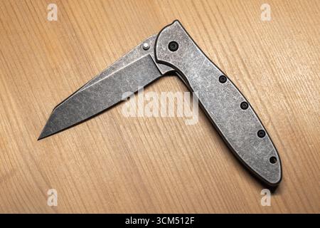 Vue détaillée d'un couteau pliant en acier gris moderne à moitié ouvert avec un design élégant reposant sur un bureau en bois Banque D'Images