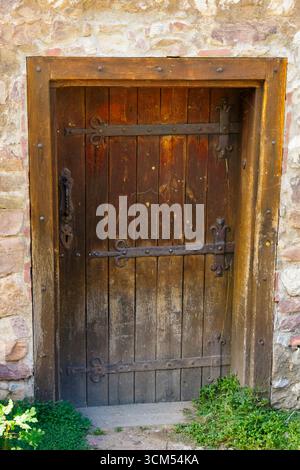 Une porte médiévale en bois robuste avec des charnières en fer ornées et une poignée, encadrée par un mur de pierre antique. L'entrée rustique reflète l'architecture de la forteresse Banque D'Images