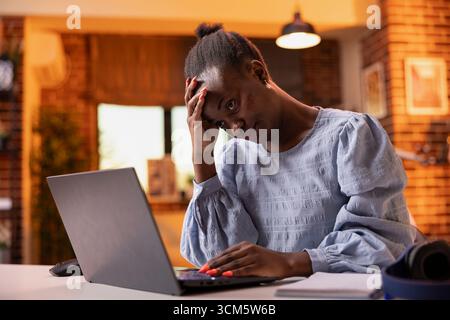 Femme d'affaires surchargée dans l'appartement, montrant la fatigue du soir pendant le travail à distance. Stressé et contrarié freelance frotte le front avec une main tout en tapant sur l'ordinateur portable, luttant pour gérer l'horaire. Banque D'Images