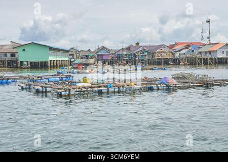 PHUKET, THAÏLANDE - 12 NOVEMBRE 2017 : village de pêcheurs côtier avec maisons sur pilotis et fermes de poissons flottants dans des eaux tranquilles sous ciel nuageux. Banque D'Images