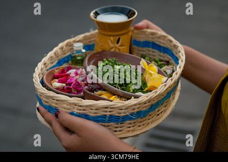 Un panier tissé rempli de fleurs colorées, d'herbes et d'une petite bouteille, tenu par une personne, avec une tasse décorative en arrière-plan Banque D'Images