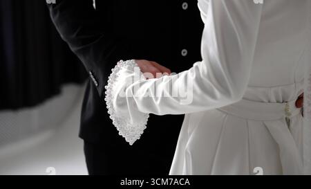 Gros plan d'un marié et d'une mariée tenant la main, présentant leur tenue de mariage et les détails de perles sur l'élégante manche blanche de la mariée Banque D'Images