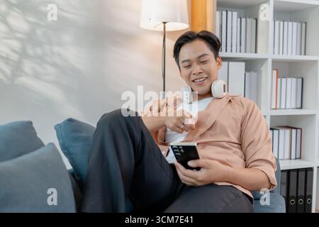 Un jeune homme se détend à la maison, souriant en utilisant son smartphone. Il aime les loisirs avec des écouteurs autour du cou, assis confortablement et feeli Banque D'Images