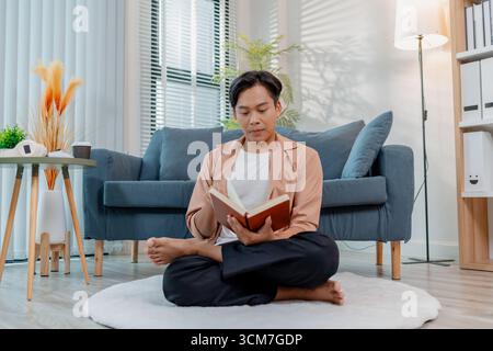 Un jeune homme est confortablement assis à l'intérieur sur le sol avec les jambes croisées, lisant un livre avec concentration et expression calme. La scène met en évidence learnin Banque D'Images
