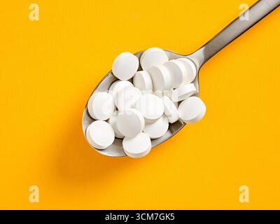 Cuillère en métal tenant de nombreuses pilules blanches rondes sur un fond jaune vif, représentant une surconsommation ou une surdose de médicaments Banque D'Images