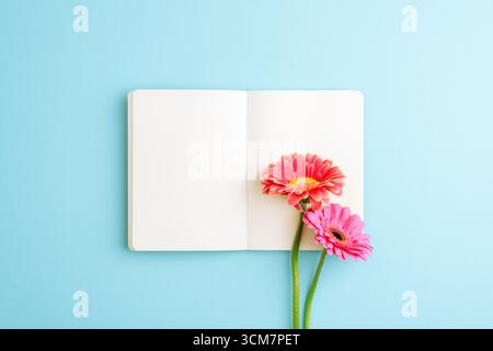 Ouvrez le carnet vierge avec des fleurs de gerbera rose et orange sur fond bleu, concept d'espace de travail créatif. Vue de dessus, pose à plat, maquette. Banque D'Images