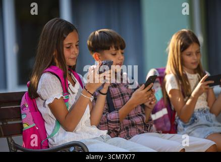 Trois enfants sont assis ensemble, concentrés sur leur smartphone, jouant à des jeux et discutant un après-midi ensoleillé après l'école Banque D'Images