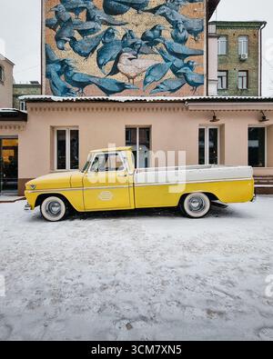 Vieux pick-up jaune garé près d'un vieux bâtiment avec murale. Vue latérale d'un camion GMC 1000 1965 dans une cour enneigée. Banque D'Images