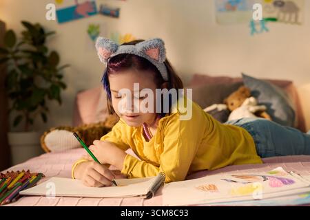 Fille caucasienne allongée sur le lit dessinant avec des crayons de couleur dans le carnet de croquis, souriant et se concentrant sur les œuvres d'art, entourée de dessins et de jouets d'animaux en peluche dans la chambre Banque D'Images