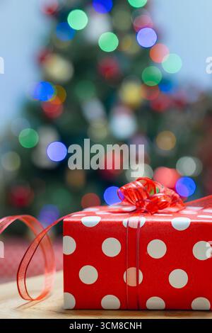 Gros plan d'un cadeau de Noël enveloppé dans du papier rouge à pois avec du ruban et des lumières bokeh festives. Ambiance de vacances, célébration, atmosphère chaleureuse Banque D'Images
