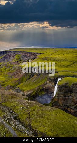 Une puissante cascade descend en cascade une falaise abrupte en Islande, entourée d'un terrain couvert de mousse, de ravins volcaniques et de lumière du soleil traversant les nuages Banque D'Images