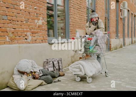 Homme caucasien sans-abri parlant au téléphone à côté d'une femme noire sans-abri qui dort Banque D'Images