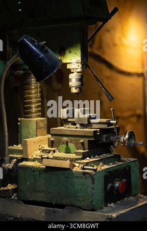 Ancienne machine à bois verte avec tête de perceuse en métal et copeaux de bois autour, éclairée par une lampe d'atelier dans un atelier de menuiserie Banque D'Images