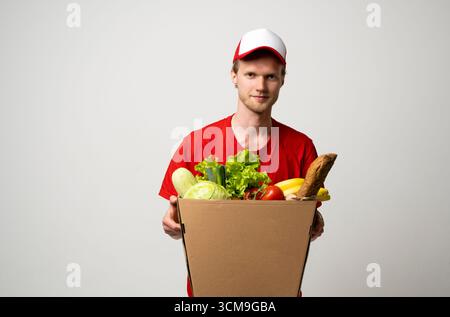 Livreur en t-shirt rouge tenant une boîte en carton avec des légumes et des fruits frais en studio Banque D'Images