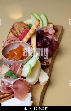 Une planche de charcuterie gourmande avec assortiment de fromages, charcuteries, raisins frais, tranches de pommes vertes, gressins, craquelins, et un bol de conserves de fruits. Banque D'Images