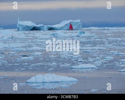 Petit voilier rouge naviguant parmi les icebergs flottants dans la mer arctique gelée sous ciel nuageux. Banque D'Images