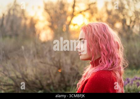 Une jeune femme aux cheveux roses éclatants se tient paisiblement dans un cadre naturel, entourée de fleurs et baignée de lumière chaude et dorée du soleil, incarnant la sére Banque D'Images