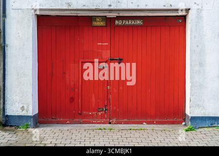 Porte rouge rustique du hangar à bateaux avec peinture pelable, panneaux d'avertissement jaunes et base pavée. Banque D'Images