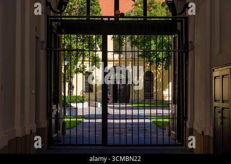 Debrecen, Hongrie 30 juillet 2025 – vue à travers la porte noire dans la cour ensoleillée avec fenêtres cintrées, toit rouge, arbres, et murs jaunes jetant du sh mou Banque D'Images
