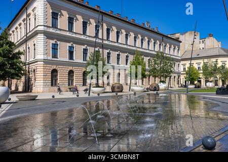Debrecen, Hongrie 30 juillet 2025 : place moderne de la fontaine d'eau avec des bâtiments historiques en toile de fond avec des tours de l'horloge et un monument de statue dans le centre-ville Banque D'Images