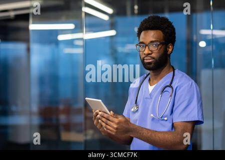 Un jeune médecin masculin portant des gommages bleus et un stéthoscope utilise une tablette numérique dans un cadre hospitalier moderne, mettant en valeur l'intersection de la technol Banque D'Images