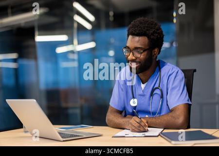 Heureux médecin afro-américain portant uniforme bleu et stéthoscope écrit sur presse-papiers tout en ayant une vidéoconférence sur ordinateur portable dans contemporain Banque D'Images