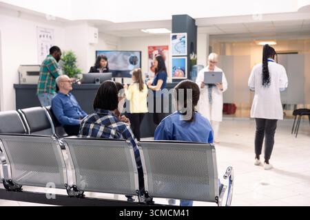 Le personnel hospitalier assiste les patients à la réception dans une salle d'attente d'urgence organisée. Infirmière dans les gommages bleus consulte l'homme asiatique assis comme d'autres patients et les médecins se préparent pour des consultations médicales. Banque D'Images