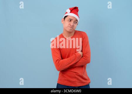 Un jeune homme réfléchi pose dans un chapeau de Père Noël festif et un pull rouge, debout contre un terrain de bal bleu vif Banque D'Images