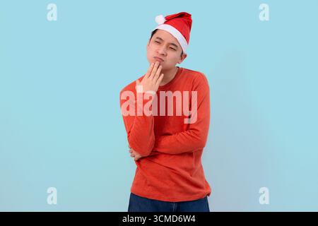 Un jeune homme réfléchi pose dans un chapeau de Père Noël festif et un pull rouge, debout contre un terrain de bal bleu vif Banque D'Images