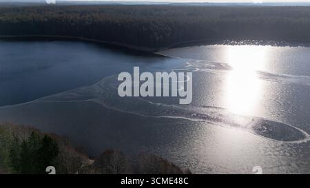 Vue aérienne d'un lac serein entouré d'une forêt dense, avec une surface partiellement gelée et une eau bleu clair. Banque D'Images