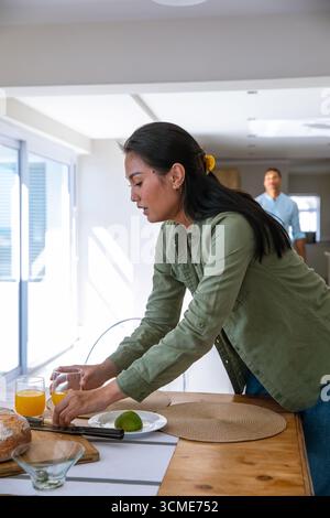 Couple diversifié arrangeant des verres de jus d'orange sur la table avec sets de table tissés, pain et citron vert Banque D'Images