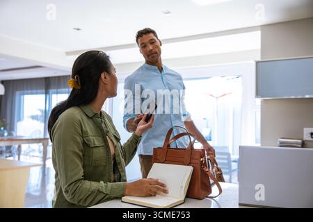 Couple diversifié échangeant smartphone à l'îlot de cuisine à côté du sac à main et ordinateur portable Banque D'Images