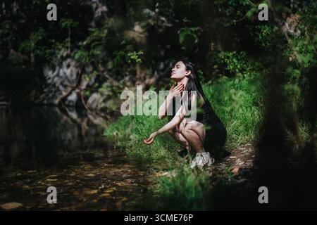 Femme agenouillée par un ruisseau forestier serein dans un environnement naturel Banque D'Images