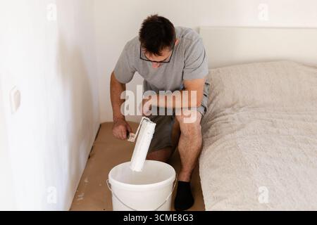 Un homme est assis sur le sol d'une chambre à coucher, peignant les murs avec un rouleau trempé dans de la peinture blanche. Il se concentre sur rendre l'espace lumineux et frais Banque D'Images