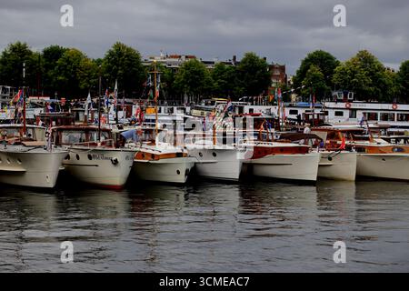 Ligne de bateaux-berlines patrimoniaux avec drapeaux et cabines en bois classiques à Sail 2025 à Amsterdam, pays-Bas, vu de l'autre côté du port avec des reflets. Banque D'Images
