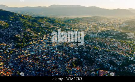 Drone vue panoramique de la ville de Sarajevo avec des toits denses, des bâtiments modernes et des montagnes lointaines dans la lumière chaude du soir Banque D'Images